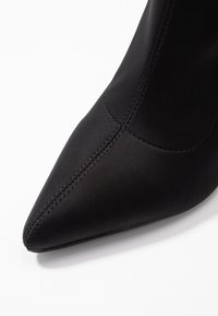 Bottine noire à talon avec un bout pointu, texture mate lisse, design épuré, et des surpiqûres visibles sur le côté.