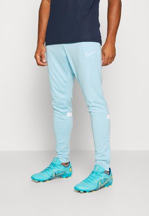 Homme portant un pantalon de sport Nike bleu clair et des crampons de football bleus assortis avec des crampons jaunes, debout sur un sol blanc avec un mur blanc en arrière-plan.