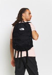 Schwarzer Rucksack mit verstellbaren Trägern, vorderem Bungee-Seil-Design und Logo. Hergestellt aus strapazierfähigem Stoff mit strukturierter Oberfläche.