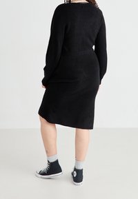 Personne portant une robe en maille noire à manches longues et des baskets montantes noires avec des lacets blancs et des chaussettes grises, debout sur un sol blanc uni.