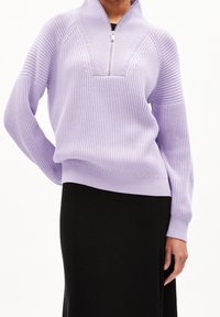 Violetti neulepullover, jossa on neljännesvetoketju, ribbitettu pinta ja leveät hihat. Yhdistetty mustaan ribbitettuun hameeseen.