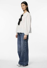 Chemisier blanc à manches bouffantes longues avec un détail de nœud noir au niveau du cou, associé à un jean bleu taille haute à jambes larges.
