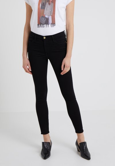 Schwarze Skinny-Jeans mit glatter Textur, hoher Taille und minimalistischem Design. Kombiniert mit einem weißen Grafik-T-Shirt und schwarzen Mules.