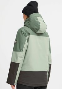 Grün und schwarz farblich abgesetzte Jacke mit Kapuze, die eine glatte Textur aufweist und ein Logo-Emblem am Ärmel hat.