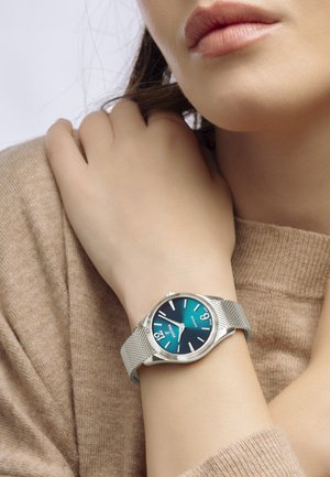 Orologio con cinturino in rete argentata presenta un quadrante verde acqua con indicatori delle ore bianchi in contrasto e un display della data alle 3. Design semplice e moderno.