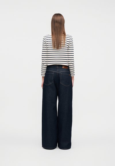 Άτομο με μακριά ίσια καστανά μαλλιά φοράει λευκό και μαύρο ριγέ πουκάμισο με μακριά μανίκια και σκούρο τζιν παντελόνι με φαρδιά μπατζάκια, στέκεται γυρισμένο προς τα πίσω.