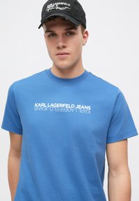 Zila kokvilnas T-kreklu ar apaļu kakla izgriezumu un īsām piedurknēm. Uzdrukāts balts vertikālais logo "KARL LAGERFELD JEANS." Gluda tekstūra.
