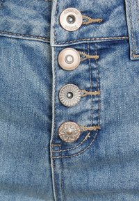 Tecido de denim azul claro com quatro botões de metal com designs decorativos, incluindo um motivo floral, padrões redondos e gravação da marca.