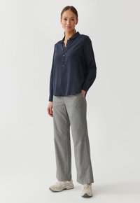 Vrouw staat, gekleed in een marineblauwe blouse met knopen, grijze broek en witte sneakers, met één hand in haar zak tegen een effen lichte achtergrond.