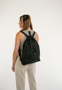 Sac à dos à cordon vert en tissu durable, avec des bretelles ajustables et un patch logo à l'avant. Texture lisse, design minimaliste.