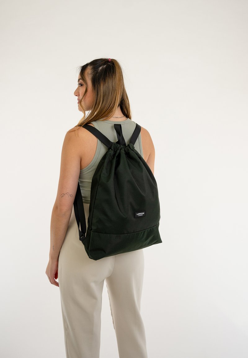 Mochila verde con cordón, hecha de un tejido resistente, que cuenta con correas ajustables y un parche con el logo en la parte frontal. Textura suave, diseño minimalista.