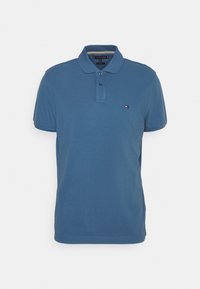 Sininen lyhythihainen polo-paita, jossa on kaksi nappia ja pieni Tommy Hilfiger -logo vasemmassa rinnassa valkoista taustaa vasten.