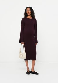 Abito premaman a coste marrone bordeaux con maniche a campana lunghe, spacco laterale e una texture liscia. Abbinato a una borsa crema e scarpe nere slip-on.