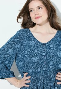 Sininen kukkakuosiset mekko, jossa on pyöreä kaulus, nappisulku ja elastinen vyötärö. Siinä on kerrostetut hihat ja pehmeä, teksturoitu kangas.