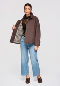 Femme portant une veste marron boutonnée avec une doublure à carreaux, un haut en dentelle noire, un jean bleu clair et des chaussures en cuir marron foncé.