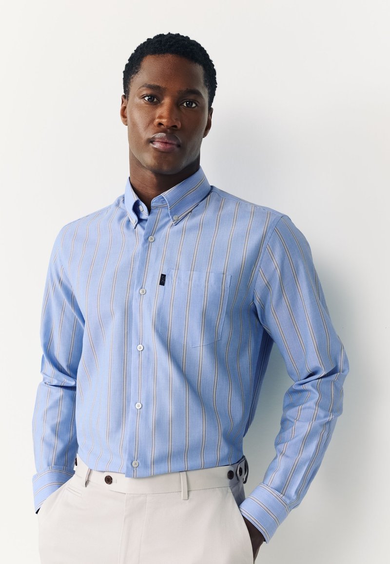 Homme portant une chemise rayée bleu clair à boutons et un pantalon beige, debout les mains dans les poches sur fond blanc.