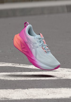 Lyseblå og lyserød løbesko i luften over asfaltvej med hvide markeringer, der viser netstof og støttende såldesign.