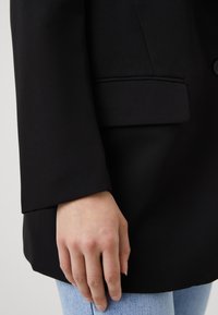 Musta bleiseri, jossa on sileä kangas, rakenteellinen muotoilu ja yksi tasku. Käsi lepää helmassa paljastaen alla vaaleansiniset farkut.