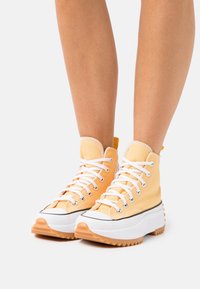 Wysokie trampki z jasno pomarańczowego płótna z białymi sznurówkami, kontrastowym czarnym wykończeniem oraz beżową, teksturowaną gumową podeszwą z wzorem w zygzak.