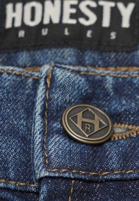 Blå denim med synlig vävstruktur, med en bronserad metallknapp med en graverad logotyp. Söm i en ljusare brun färg.
