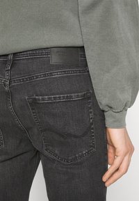 Black denim jeans with a smooth texture, featuring a leather patch labeled "JACK & JONES" and a classic five-pocket design.