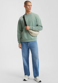 Uomo che indossa un felpa verde chiaro, jeans blu, sneakers bianche e una borsa a tracolla beige, in piedi davanti a uno sfondo grigio uniforme.