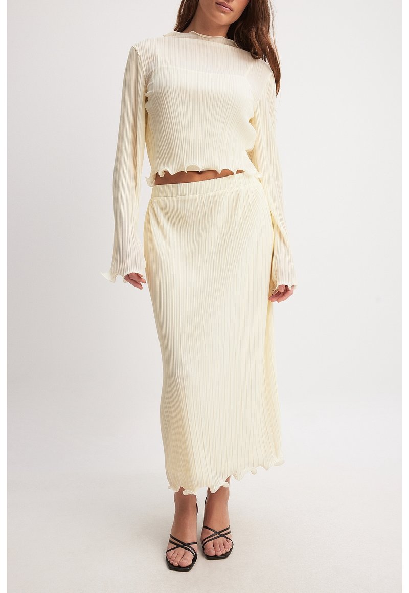 Femme portant un crop top côtelé crème à manches longues et une jupe midi assortie avec des bords ondulés et des sandales noires à brides.