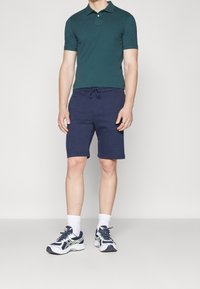 Zelené polo tričko s límečkem, doplněné o tmavě modré šortky na šňůrku. Bílé sportovní ponožky a tmavě modré/bílé tenisky dotvářejí celý outfit.