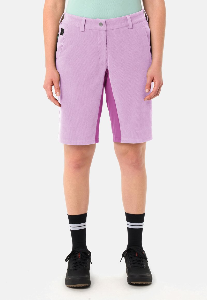 Personne portant un short en velours côtelé violet clair atteignant les genoux, un haut vert menthe, des chaussures de sport noires et des chaussettes noires avec des rayures blanches, debout devant un fond blanc.