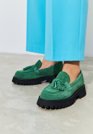 Grüne Wildlederloafers mit einer klobigen schwarzen Sohle, die an der Vorderseite mit einem dekorativen Quasten verziert sind und eine glatte Textur aufweisen. Kombiniert mit hellblauen Hosen.
