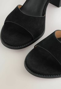 Black suede sandals with a low block heel, featuring crossed straps over the toes and a smooth inner lining, resting on a light background.