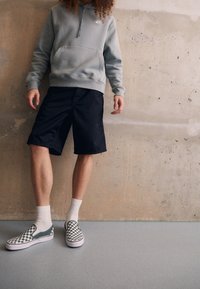 Grå hoodie med en framficka, svarta shorts och grön-och-vit rutiga slip-on skor, kombinerat med vita ankelsockor.