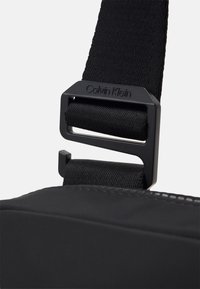 Cintura a spalla in nylon nero con motivo testurizzato, dotata di una fibbia in metallo nero opaco riportante la scritta "Calvin Klein." Forma rettangolare.