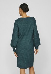 Donna con i capelli raccolti in un chignon, che indossa un vestito a maniche lunghe in maglia color teal scuro, in piedi con la schiena rivolta verso la macchina fotografica su uno sfondo neutro.