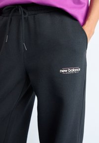 Gros plan sur une personne portant un pantalon de survêtement noir New Balance avec taille à cordon et un haut violet, main dans la poche droite.