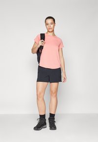 Rosa kortermet t-skjorte, parret med svarte shorts. Modellen har på seg svarte turstøvler og bærer en svart ryggsekk over den ene skulderen.