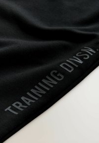 Schwarzer Stoff mit einer glatten Textur zeigt den eingeprägten Text "TRAINING DIVSN." in einem glänzenden Finish entlang der Kante.