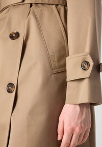 Khaki płaszcz trench z gładką teksturą, podwójną zapięciem, okrągłymi guzikami i widocznym logo na bocznej kieszeni.