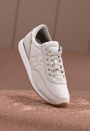 Jasnobeżowy sneaker Saucony z sznurowadłami, panelami z zamszu i siatki, białą środkową podeszwą i gumową podeszwą zewnętrzną na miękkiej brązowej powierzchni.
