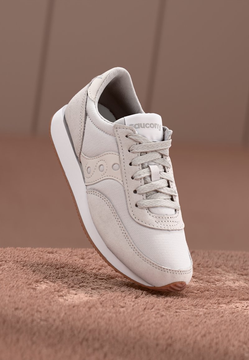 Baskets Saucony beige clair avec lacets, empiècements en daim et mesh, semelle intermédiaire blanche et semelle extérieure en gomme naturelle sur une surface marron douce.