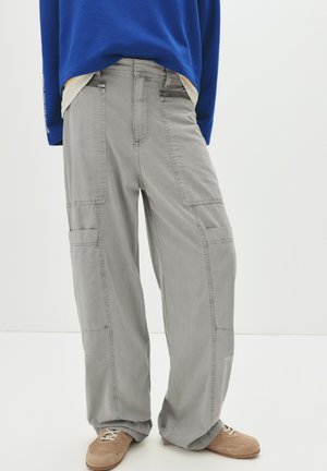 Pantaloni cargo grigi dal taglio rilassato, con molteplici tasche e un tessuto liscio e strutturato. Abbinati a una camicia a maniche lunghe blu.