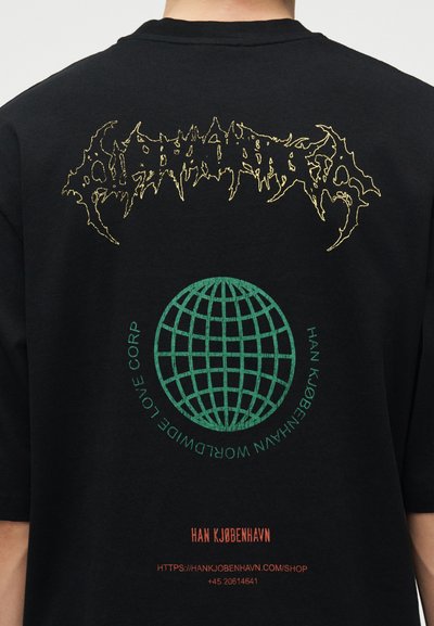 Άτομο που φοράει μαύρο πουκάμισο με κίτρινο αφηρημένο κείμενο στο πάνω μέρος, πράσινο γραφικό γήινου στο κέντρο και κόκκινο όνομα εμπορικού σήματος και κείμενο ιστοσελίδας κάτω από αυτό.