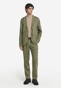 Grüner Leinenanzug mit einreihigem Blazer, Reverskragen und zwei Knöpfen. Kombiniert mit einem beigen Hemd und dunklen Schuhen. Glatte Textur.