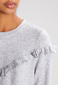 Maglione grigio con scollatura rotonda decorato con un accento arricciato lungo una spalla. Il tessuto ha un aspetto morbido e testurizzato.