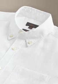 Chemise blanche boutonnée avec col boutonné, poche avant et étiquette de marque visible à l'intérieur du col.