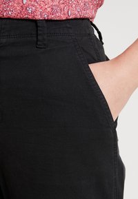 Pantalons en mélange de coton noir avec une texture lisse, dotés de poches latérales et d'une taille mi-haute, partiellement visibles contre un haut à motifs.