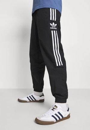 Zwarte sportbroek met het witte Adidas-logo en drie verticale strepen aan elke kant, gecombineerd met witte sneakers met marineblauwe accenten.