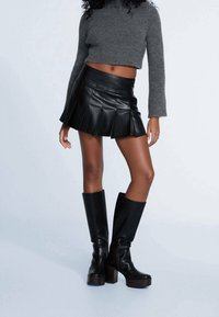 Schwarzer, plissierter Minirock aus Leder, kombiniert mit kniehohen schwarzen High-Heels Stiefeln. Das Outfit wird durch einen cropped grauen Pullover ergänzt.