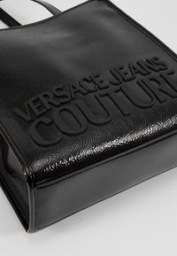 Czarna torebka o fakturze z wytłoczonym logo "Versace Jeans Couture" z przodu, przedstawiona na gładkim jasnym tle.