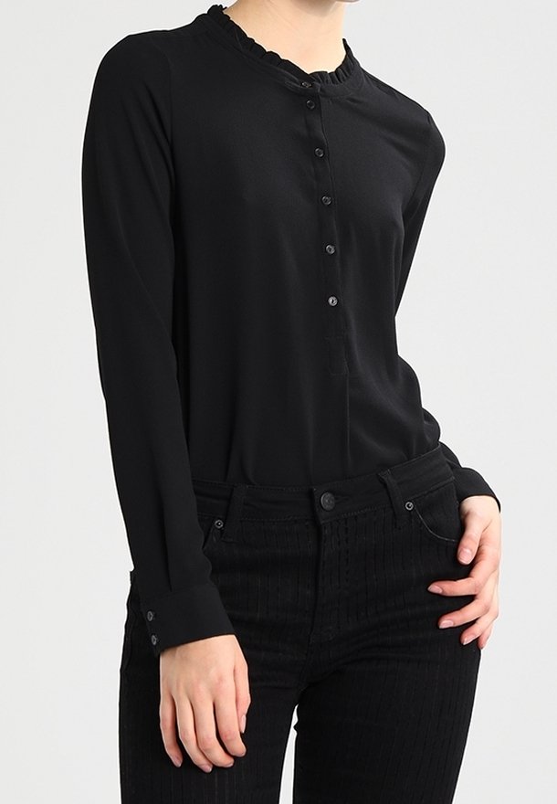 Blusa nera a maniche lunghe con bottoni e colletto arricciato indossata con pantaloni neri a vita alta, una mano appoggiata sui fianchi, sfondo neutro.